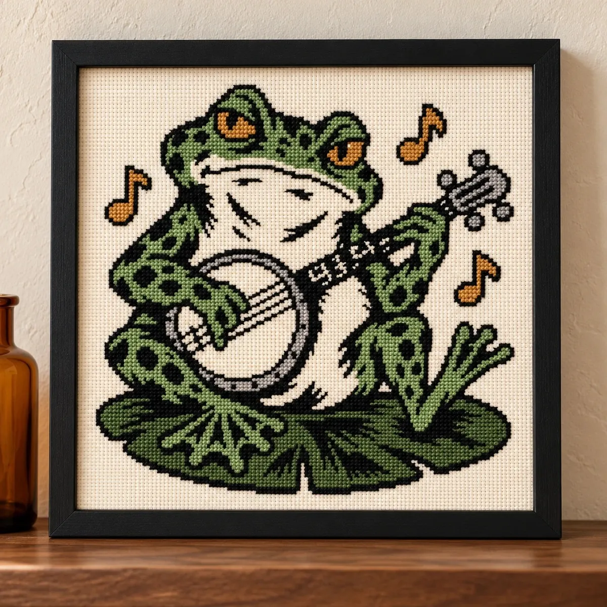Straight-on view of the finished Frog Playing Banjo Cross Stitch in a black frame on a wooden shelf