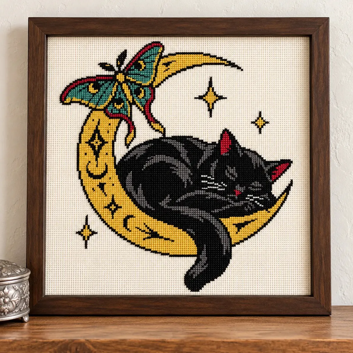 Black cat sleeping on a gold crescent moon with a luna moth, cross stitch in a brown frame on a shelf