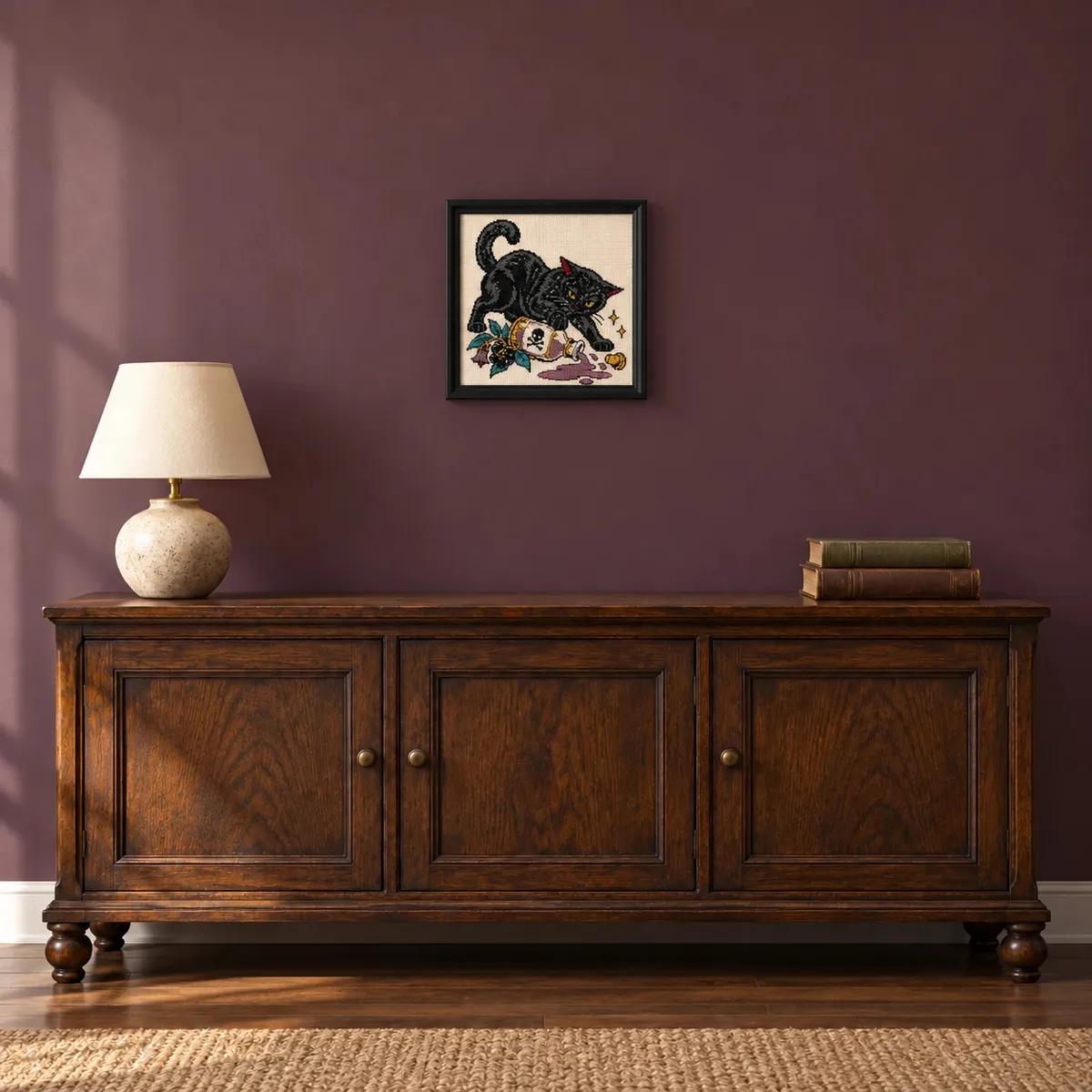 Framed black cat and spilled poison cross stitch on a plum wall above a wood cabinet