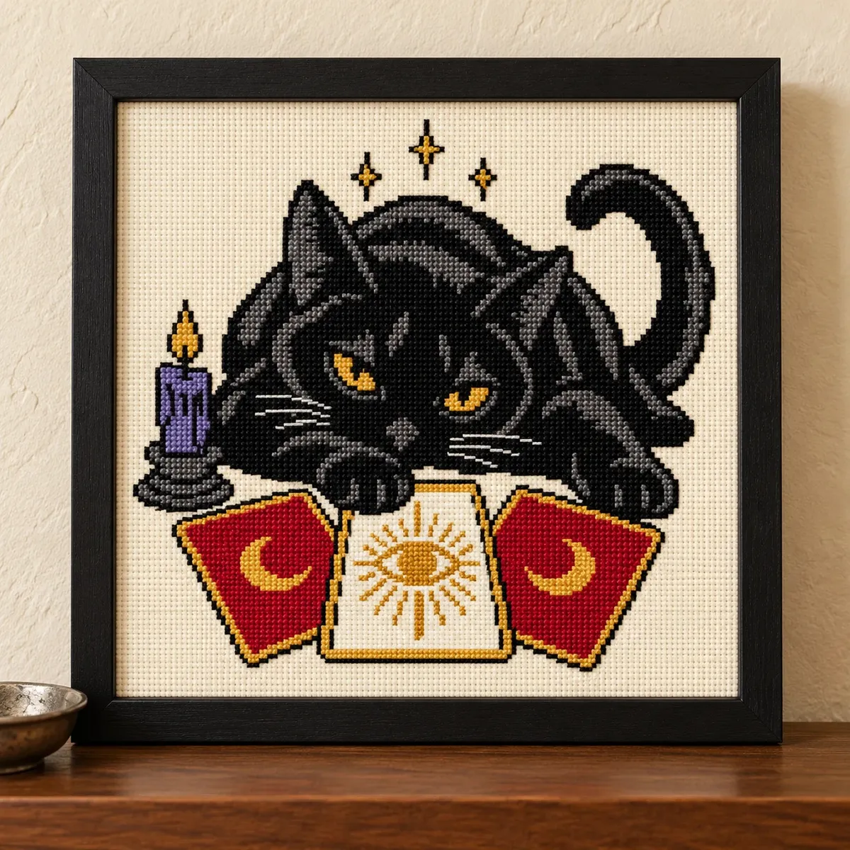 Black cat lying over three tarot cards, cross stitch in a black frame on a wood shelf